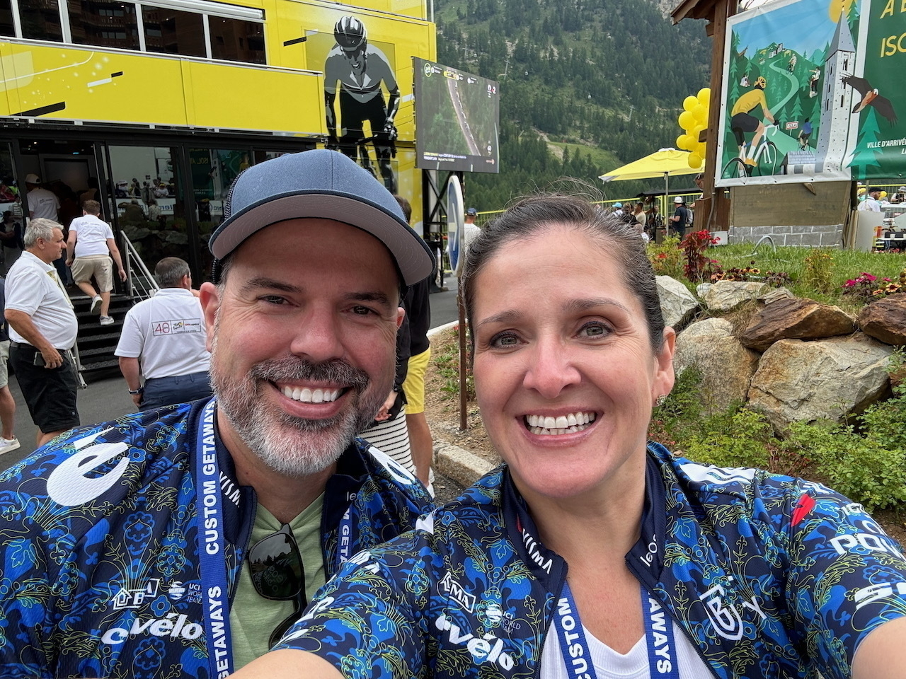 Auto-generated description: Two people are taking a selfie outdoors, wearing matching cycling jerseys with a mountain scene and a yellow building in the background.