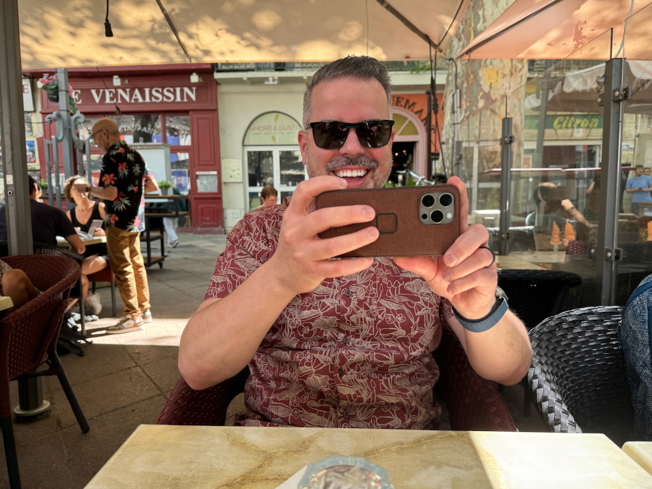 Auto-generated description: A man with sunglasses is smiling and holding a smartphone in an outdoor café setting.